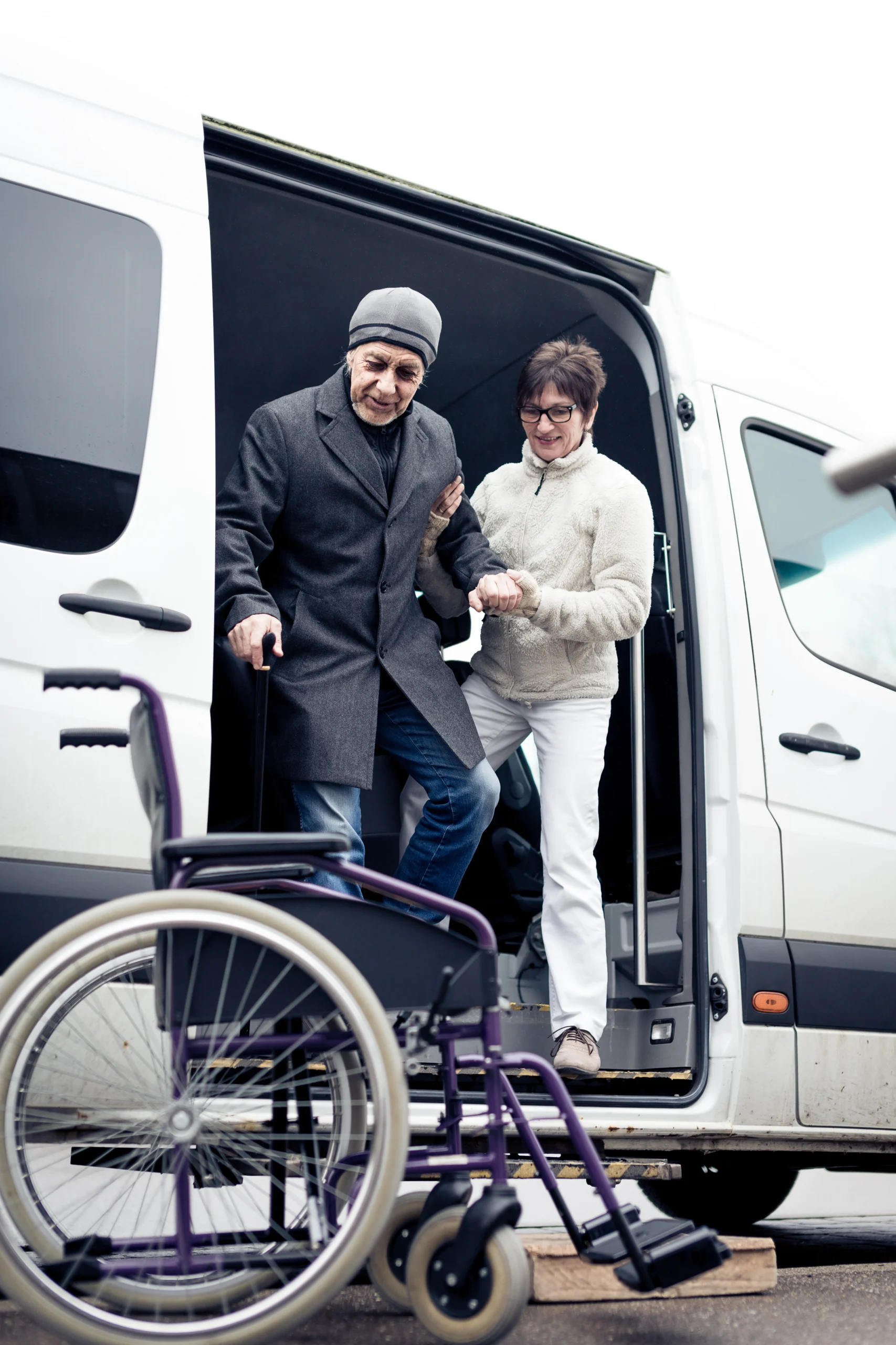 Caregiver helping an elderly man with a cane step down from a medical vehicle for ambulatory transport.