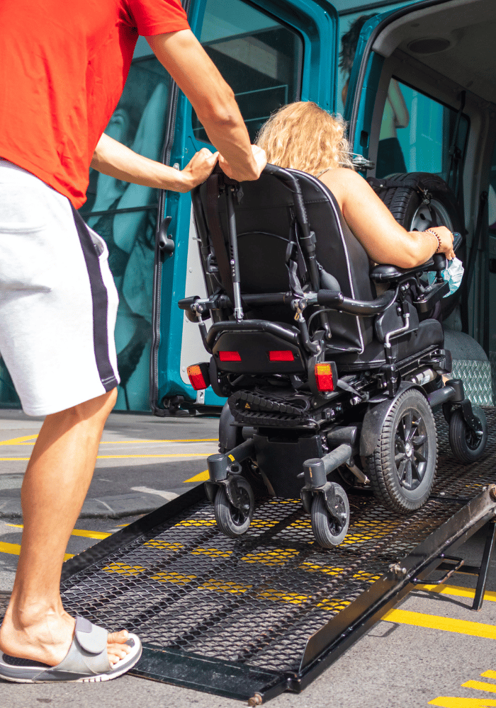 Caregiver pushing a passenger in a motorized wheelchair up a ramp into an ADA accessible medical vehicle.